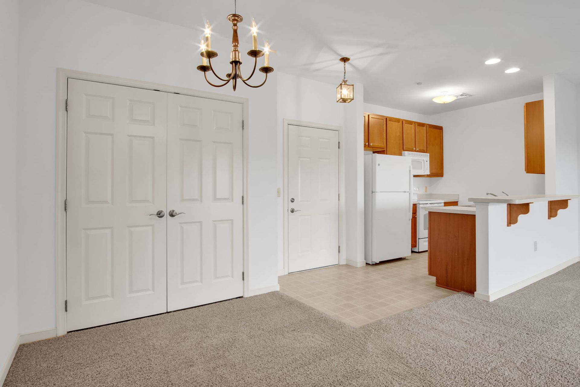 Empty carpeted room with chandelier, white double closet doors, and a small kitchen area with wood cabinets.