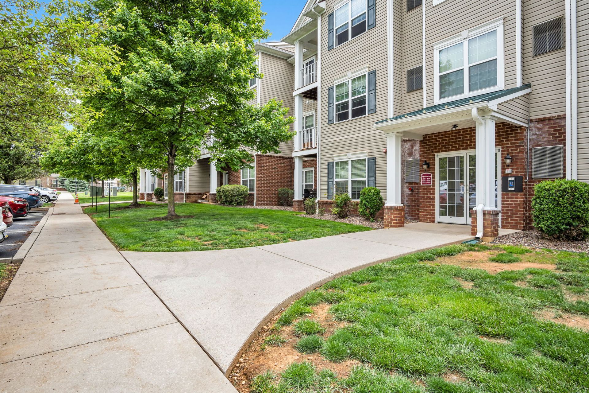 Apartment building entrance with sidewalk, green lawn, and leafy tree
