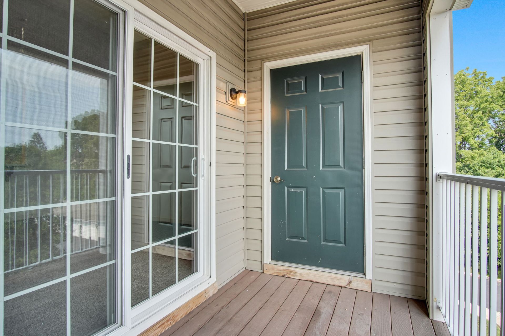 Apartment balcony with teal front door, white siding, and large windows beside a wooden deck.