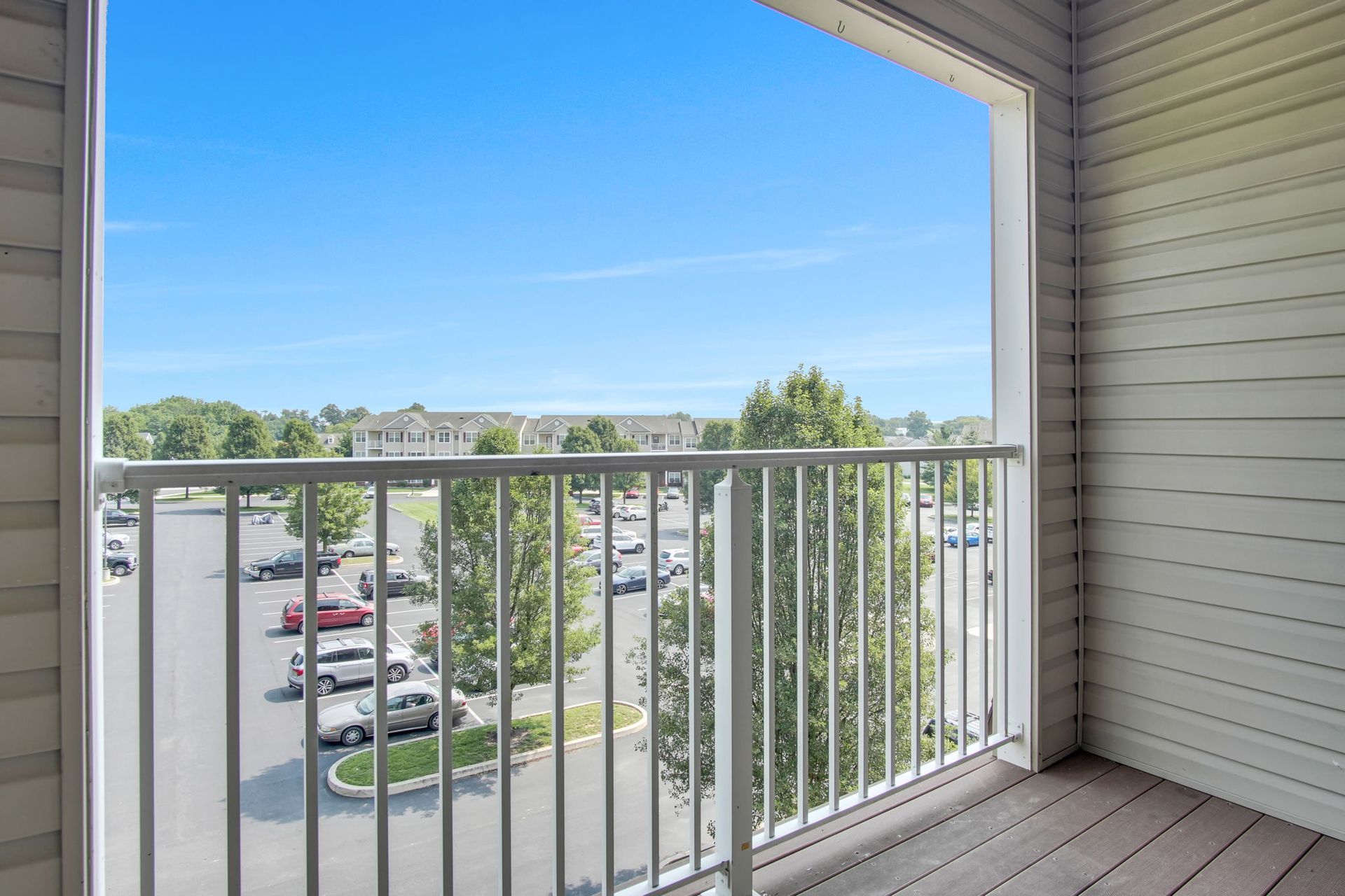 Covered balcony with white railing overlooking a parking lot and trees under a blue sky