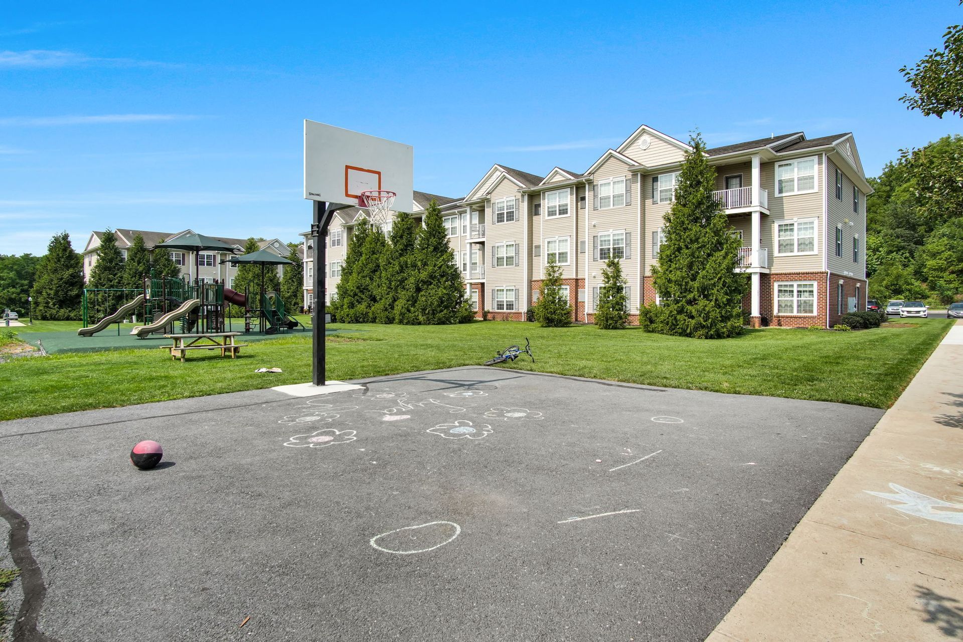 Outdoor basketball court beside an apartment building on a sunny day, with a ball near the hoop.