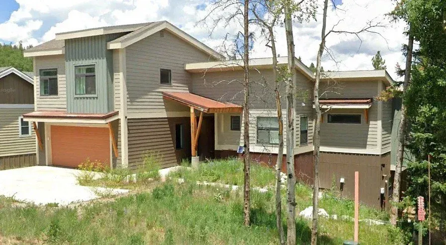 Two-story gray house with red brick accents, a front porch, and tall trees in a grassy yard