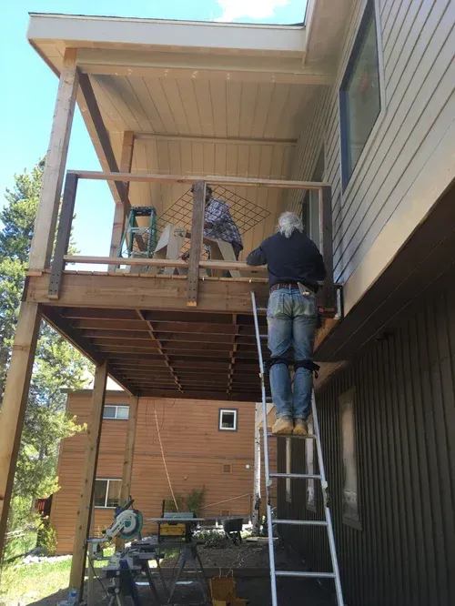 Person on ladder working on a wooden balcony/porch exterior beside a house.