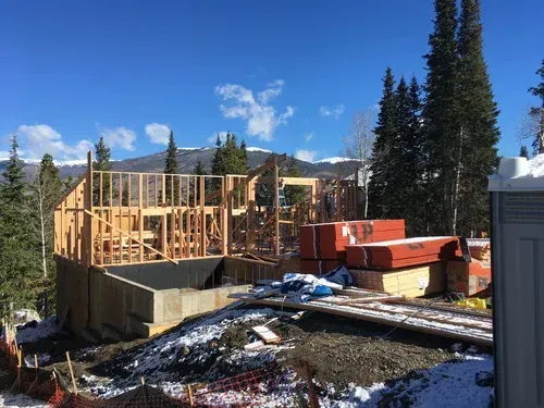 Wood-framed house under construction with red dumpster and snow on the ground under a bright blue sky