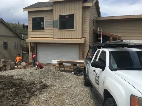 House under construction with workers, gravel driveway, and a white pickup truck in front