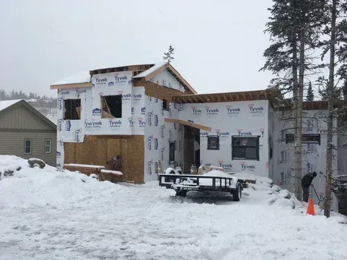 Snow-covered house under construction with exposed wood framing and wrapped walls in a winter setting