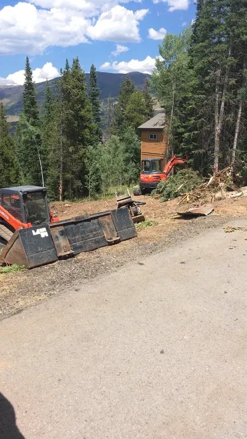 Construction equipment on a dirt road in a mountain forest with a cabin and blue sky.