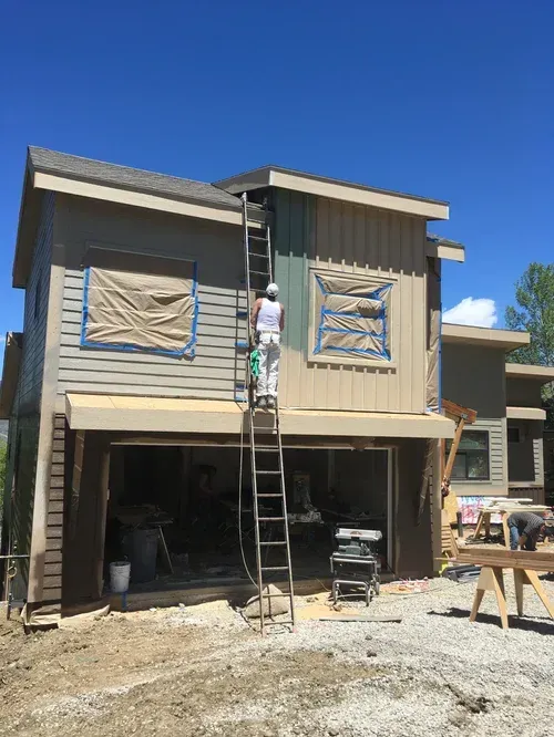 Worker painting second-story exterior of a house under construction, with windows taped off and ladder below.