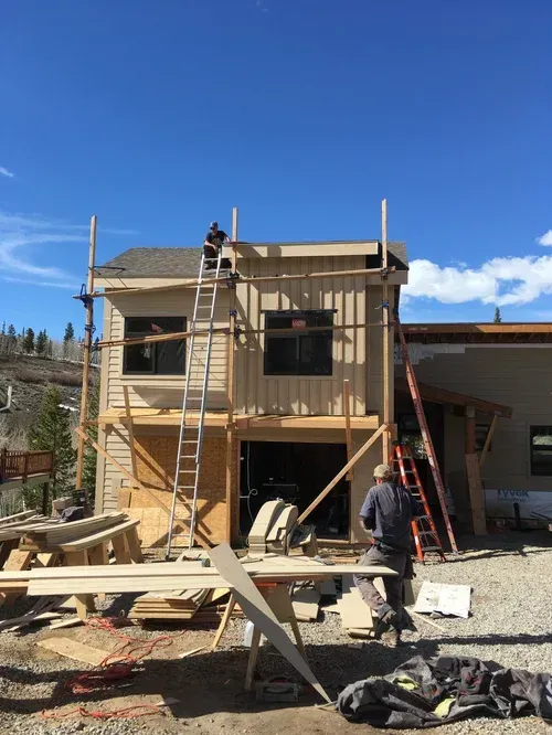 Two workers on ladders building a small wooden house under a clear blue sky