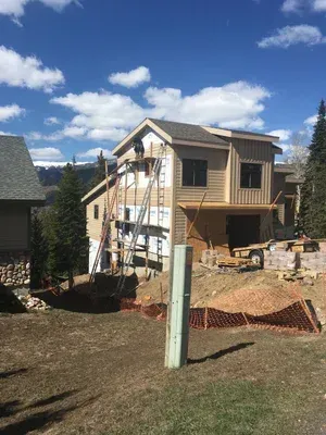 House under construction with ladders, scaffolding, and orange safety fencing in a grassy yard under a blue sky