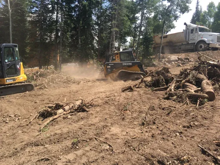 Construction equipment clearing a dirt lot with tree stumps and a dump truck in the background.
