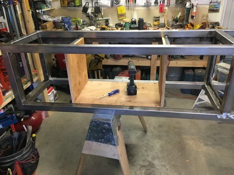 Metal frame on a workbench in a garage, with a drill standing in the center.