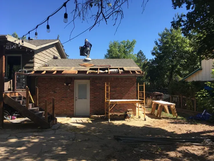 Backyard shed under construction with a worker on the roof and lumber stacked beside it