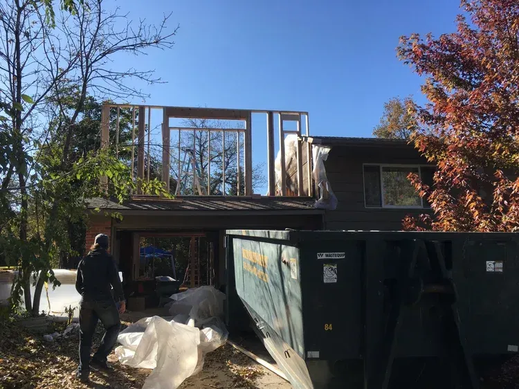 Construction site with dumpster, worker, and partially built house frame under clear sky