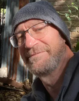 Man in a gray beanie and glasses smiling outdoors in sunlight