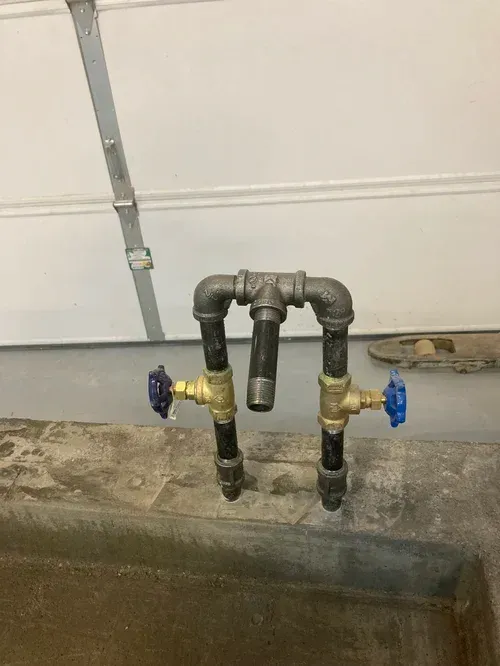 Two black pipes with brass and blue shutoff valves against a garage wall.