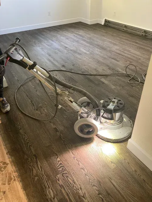 Floor buffer sanding a wooden floor in an empty room with gray walls.