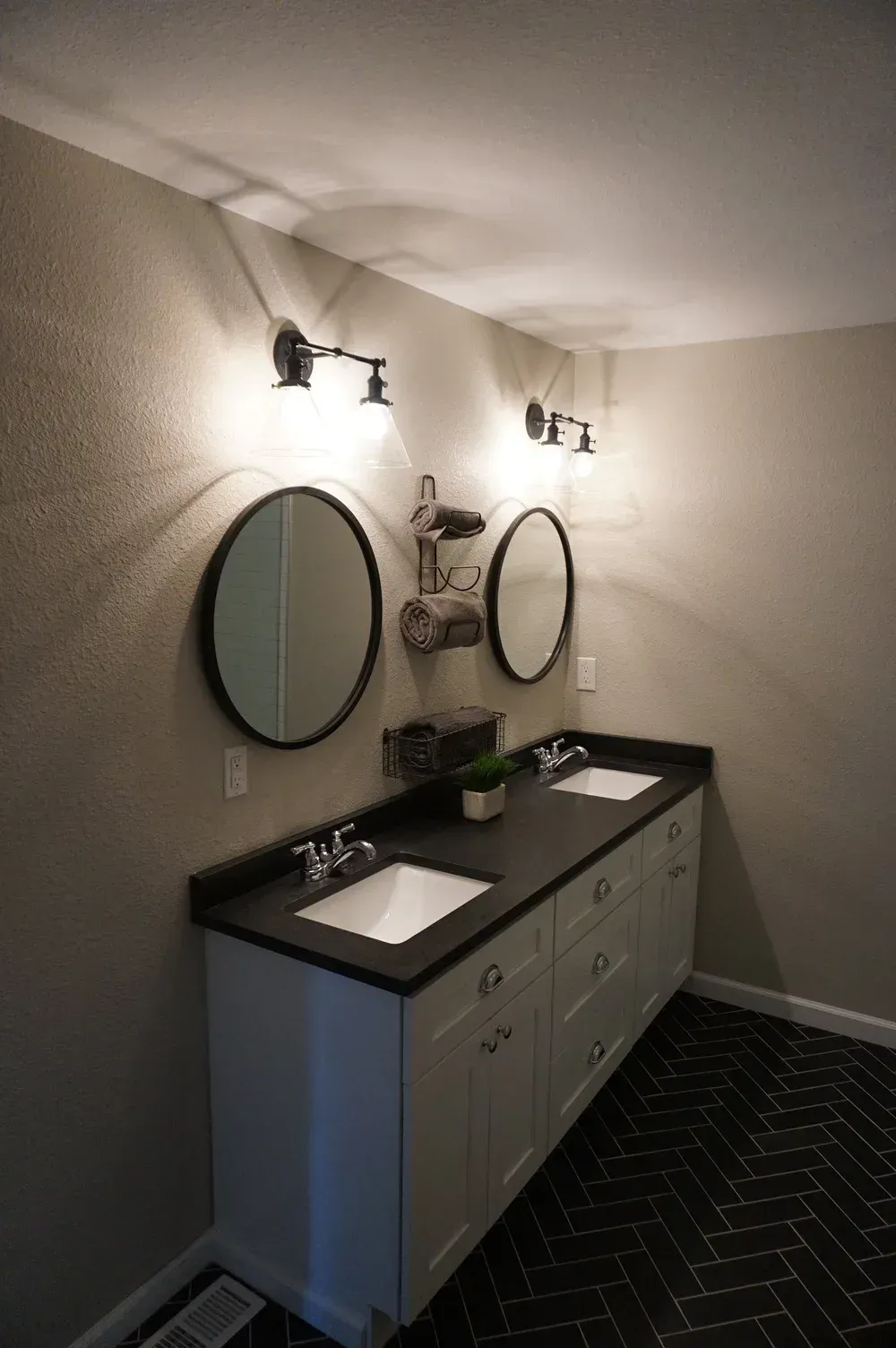 Modern bathroom with double sinks, round mirrors, black countertops, and wall sconces on beige walls.