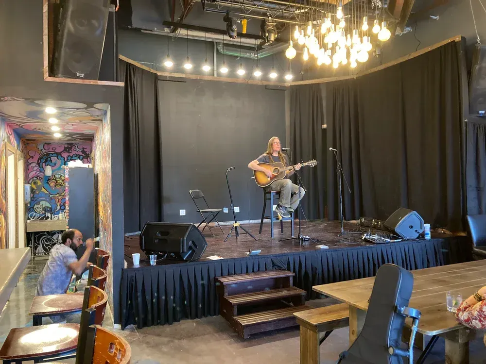 Small stage performance in a cozy venue, with a guitarist under string lights and black curtains.