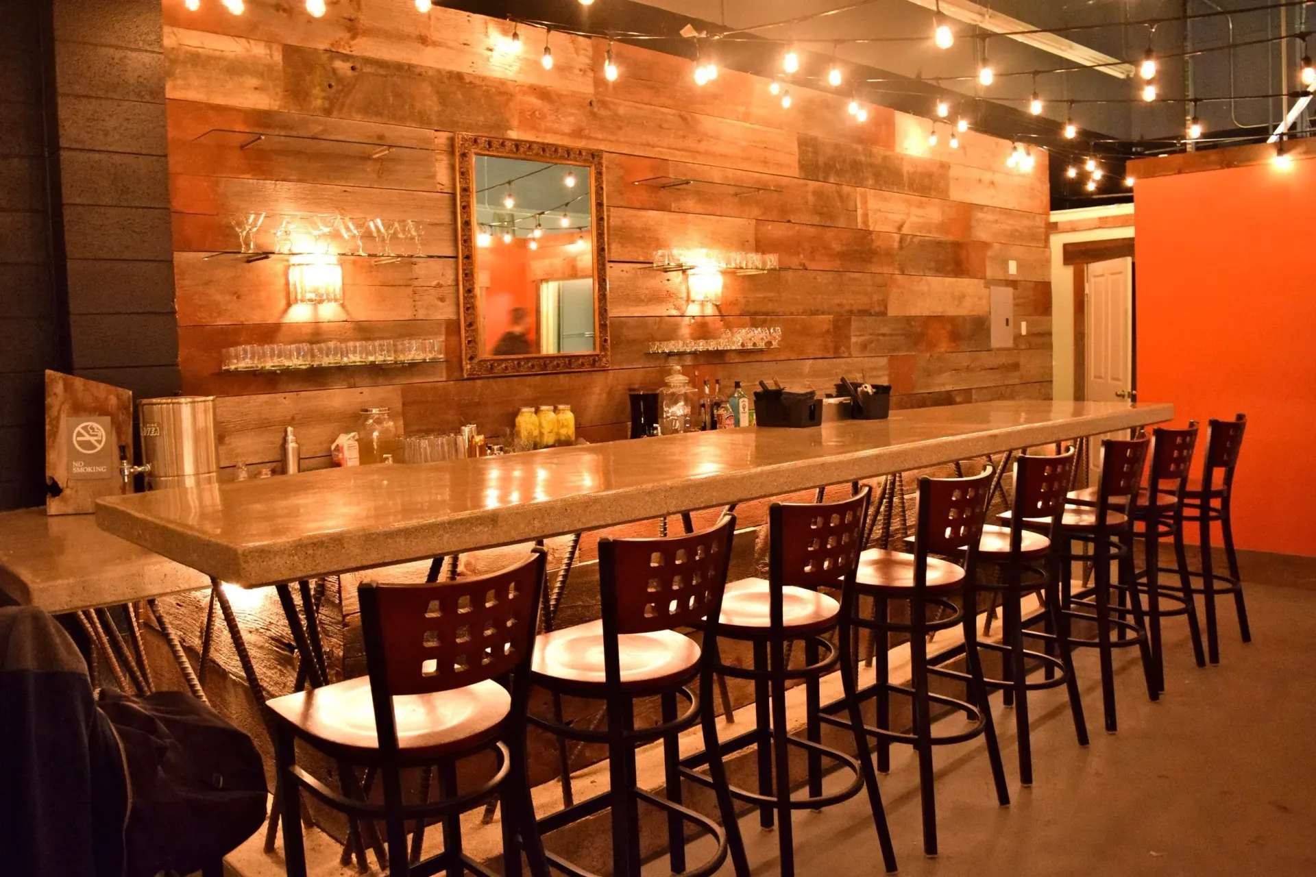 Warmly lit bar with long wooden counter, stools, brick wall, and hanging string lights