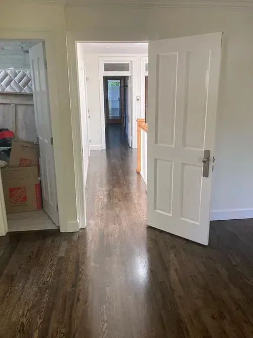 Empty hallway with dark wood floors, white walls, and open doors leading to another room.