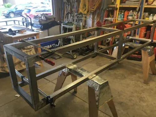 Metal rectangular frame being welded in a cluttered workshop garage with tools and shelves nearby