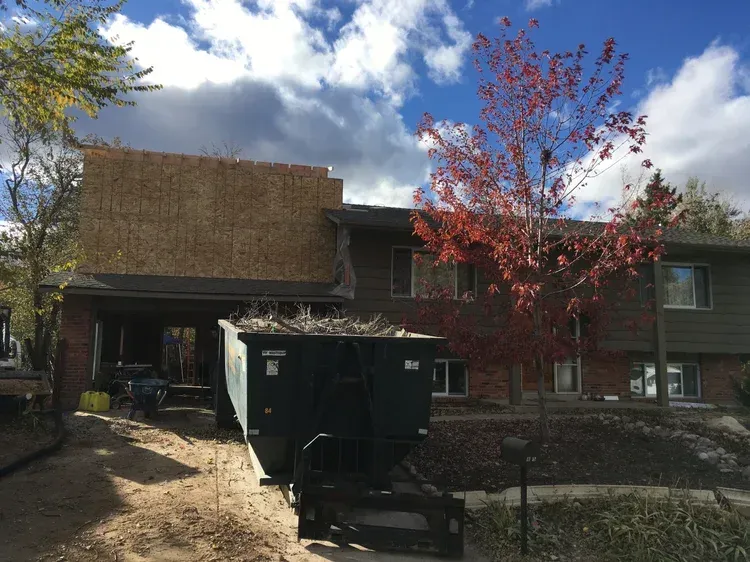 Residential building with brick facade, black dumpster in front, and a red-leaved tree in the yard