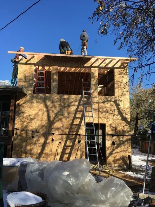 Two workers on a roof framing a second story of a house with ladders and construction materials below