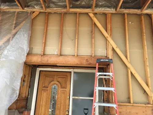 Interior wall under renovation with exposed studs, insulation, and a ladder beside a front door.