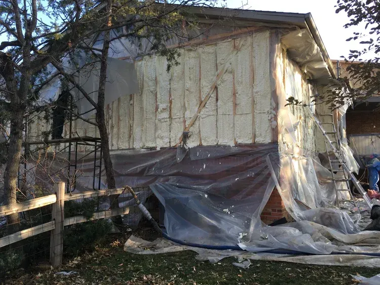 House wrapped in plastic sheeting during exterior renovation, with ladder and debris around it
