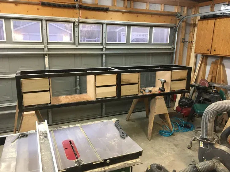 Workbench frame under construction in a garage, with tools and wood panels on a sawhorse.