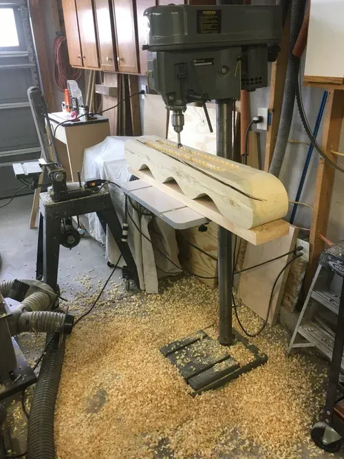 Workshop drill press shaping a large wood piece on a stand, with sawdust scattered on the floor.