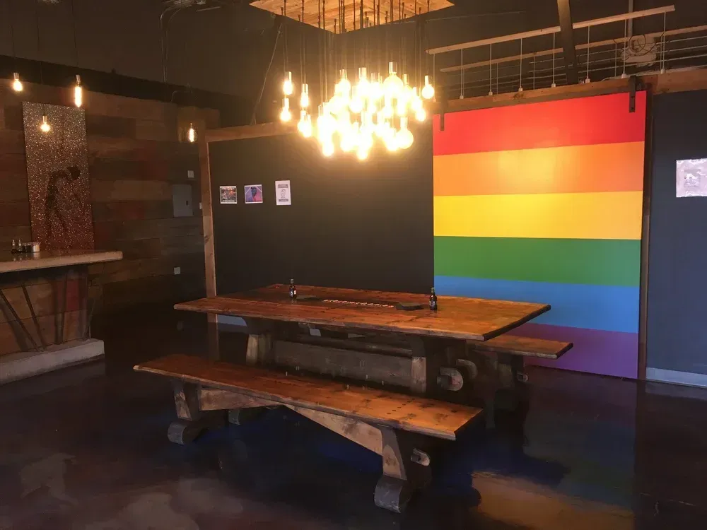 Rustic dining room with wooden tables, chandelier, and a large rainbow pride flag on the wall