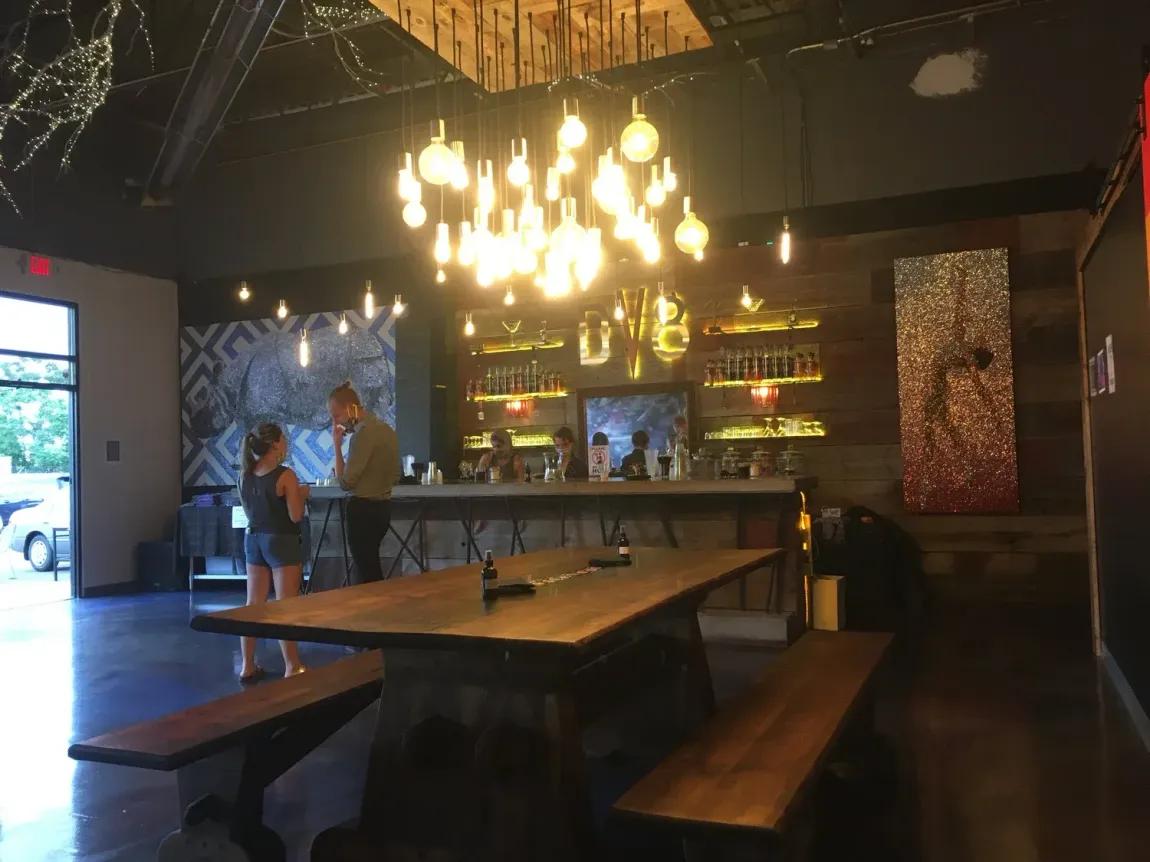 Dim restaurant interior with long wooden tables, benches, hanging lights, and a bar in the background