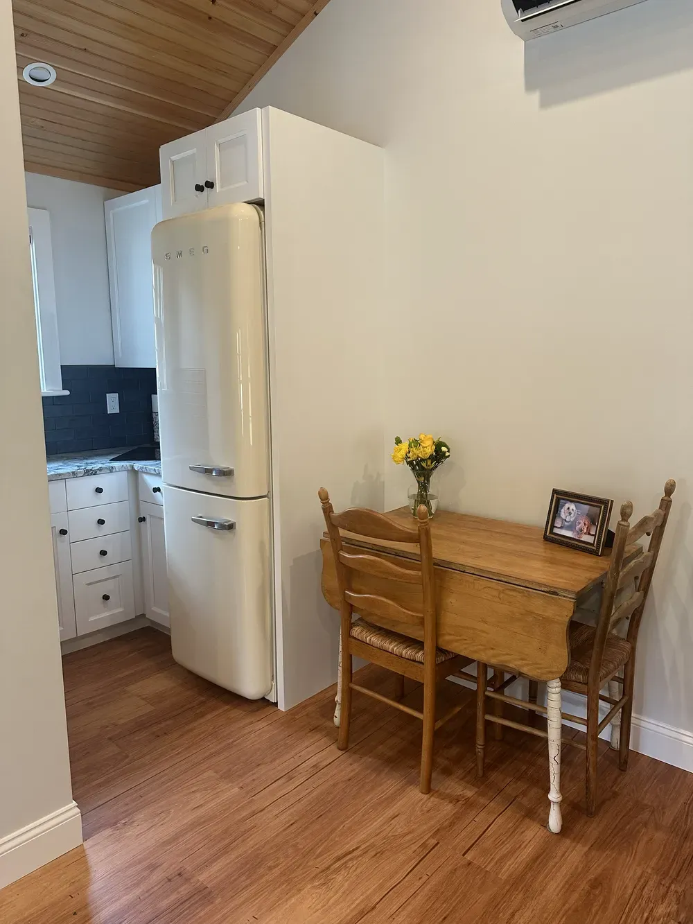 Small kitchen nook with a white fridge, wooden table and chairs, and a vase of yellow flowers near a window.