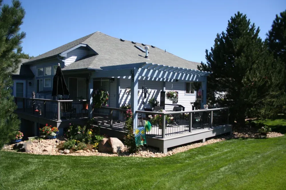 Gray house with a deck and pergola, surrounded by green lawn and trees under a clear blue sky