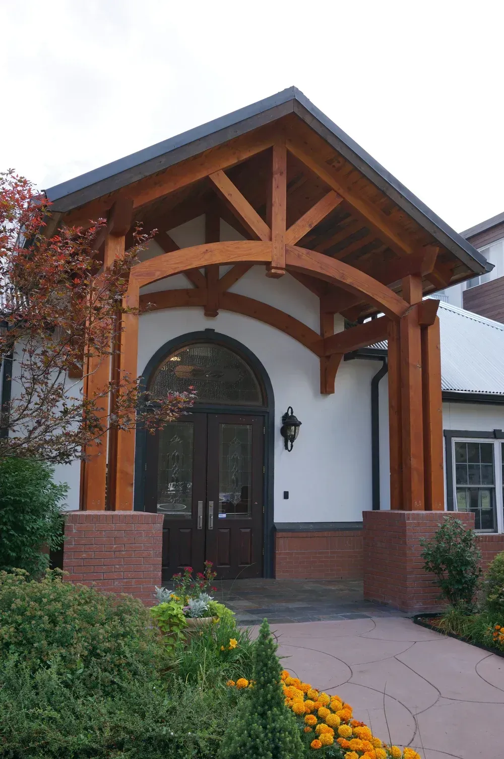 Front entrance with timber canopy, arched glass door, white stucco walls, and flower beds along a stone path