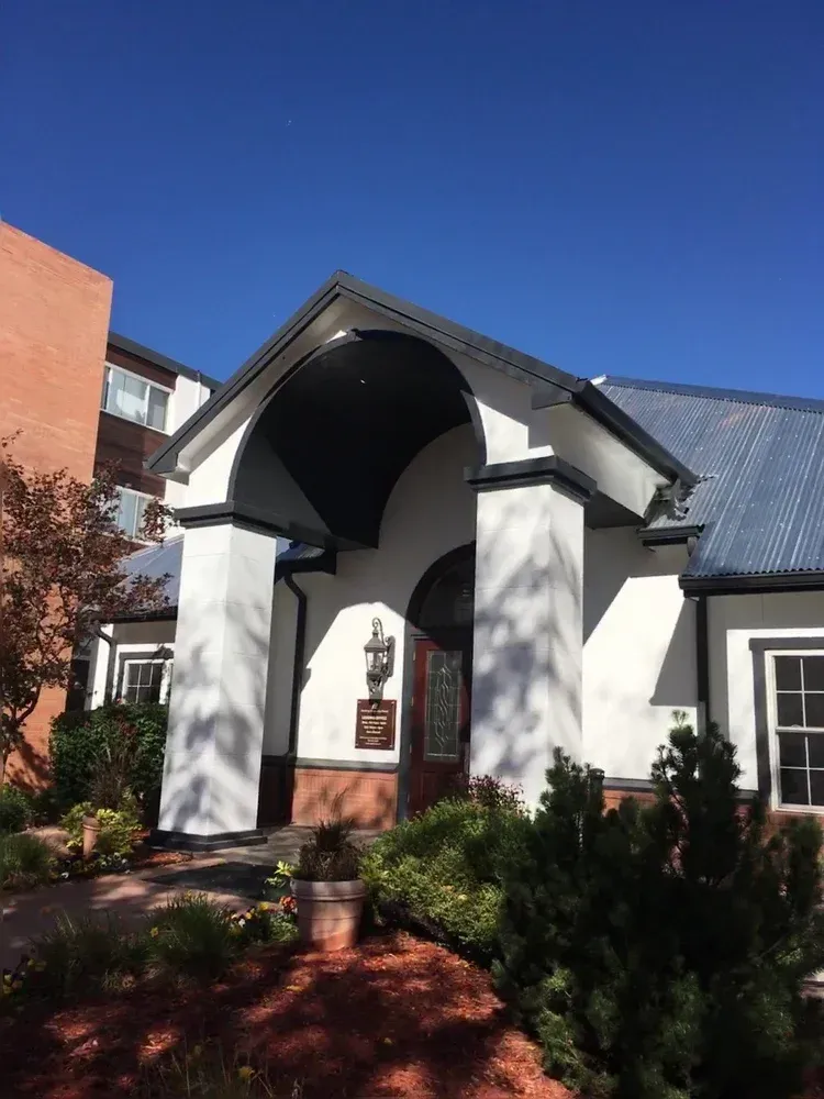 White church entrance with arched portico, dark roof, and landscaped shrubs under a clear blue sky.
