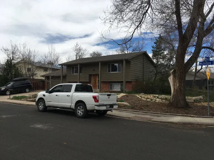 White pickup truck parked on a residential street in front of a green house with bare trees.
