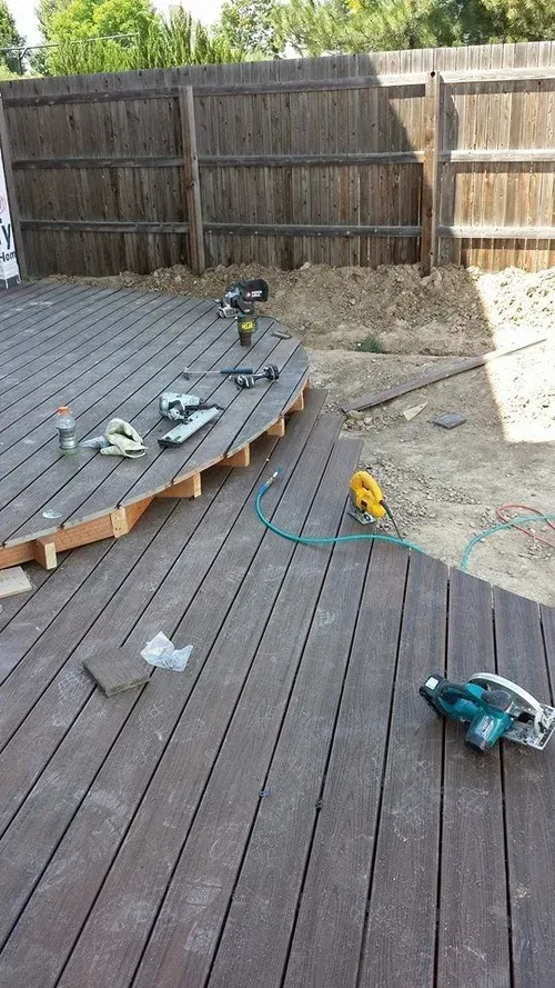 Backyard deck under construction with tools, lumber, and a circular raised platform by a wooden fence
