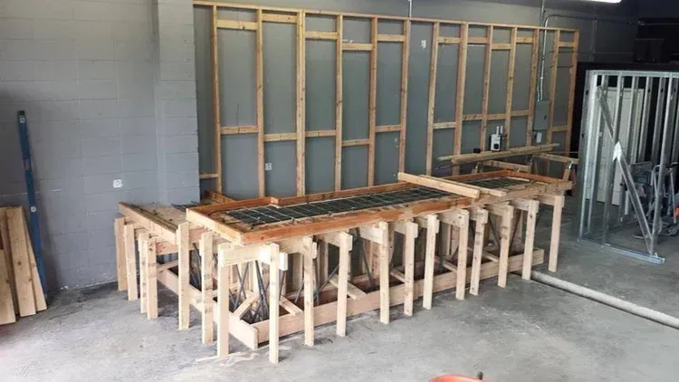 Wood-framed construction project in a garage, with stacked lumber and wall framing against a gray wall