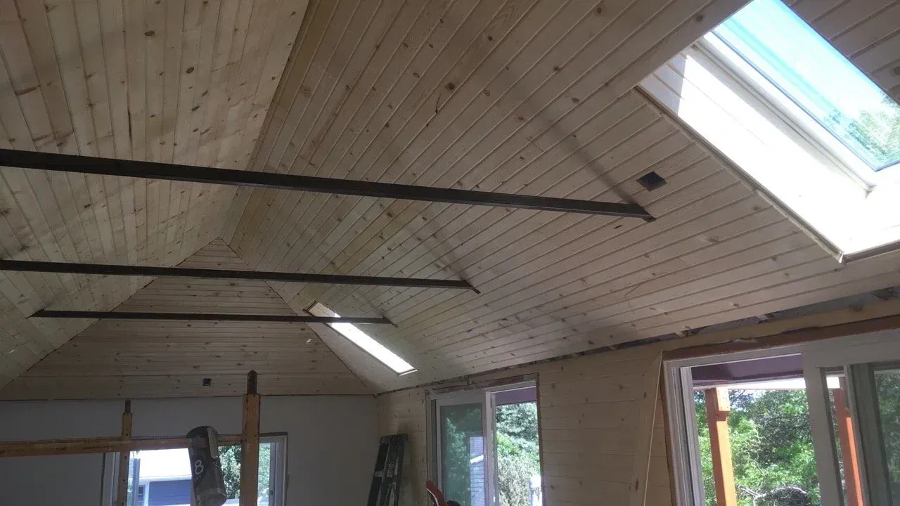 Interior of a room with a white wood-paneled vaulted ceiling, skylights, and large windows/doors below.