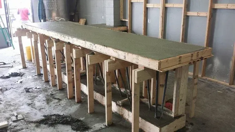 Long wooden workbench under construction in a workshop, with exposed supports and a concrete top.