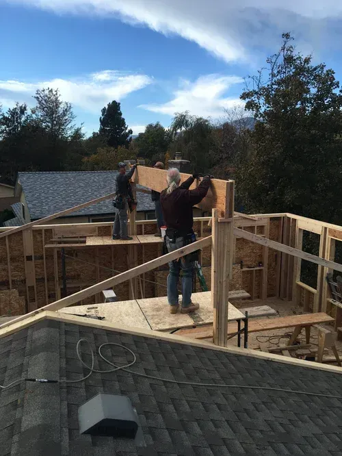 Person on a rooftop under construction, lifting lumber amid wooden framing and cloudy sky