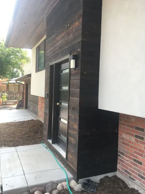 Exterior view of a modern doorway with dark wood siding, brick wall, and a green hose on the ground