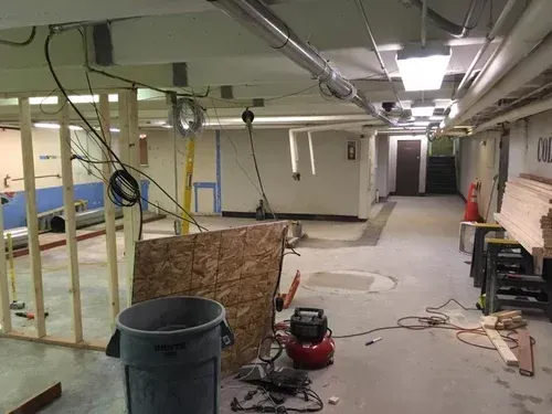 Basement under renovation with exposed pipes, debris, tools, and a trash can on the floor.