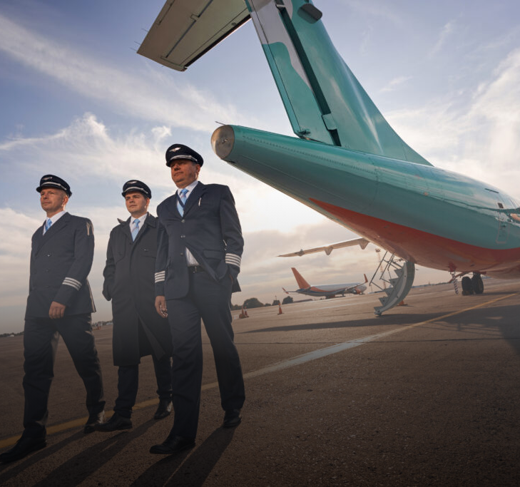 Three pilots in uniform stand near a teal airplane on an airport tarmac.