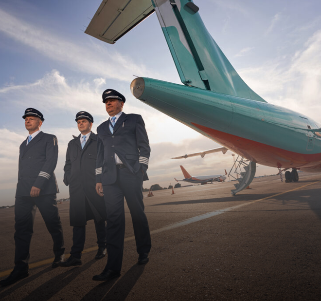 Three pilots in uniform stand on tarmac near a teal airplane, another plane in the distance.