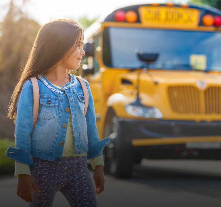 Girl with a backpack, wearing a denim jacket, looks towards a yellow school bus.