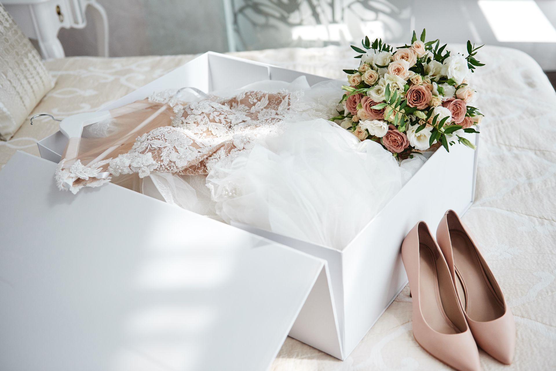 Bride’s white dress, nude heels, and pastel bouquet in an open white box on a bed